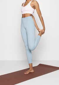 Sujetador deportivo rosa claro y leggings de cintura alta azul claro, ambos ajustados. La modelo realiza una postura de yoga sobre una esterilla marrón.