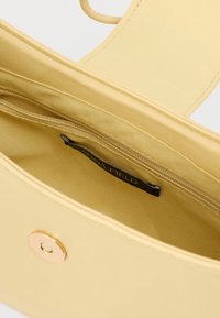 Κίτρινη γυναικεία τσάντα ανοιχτή για να δείξει την εσωτερική τσέπη με φερμουάρ, με μαύρη ετικέτα και χρυσή μαγνητική κλειδαριά στην μπροστινή πτέρυγα.