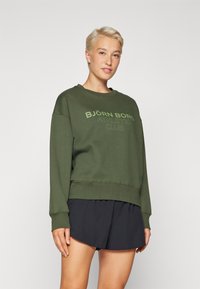 Olívazöld pulóver, kerek nyakkal, oversized fazonban, világoszöld „BJÖRN BORG ATHLETIC CLUB” logóval. Fekete rövidnadrággal párosítva.