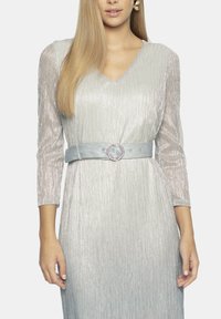 Srebrna sukienka z długim rękawem w kształcie litery V, z błyszczącymi pionowymi paskami, paskiem w talii oraz jasnoniebieskim paskiem z okrągłym detalem metalowym.