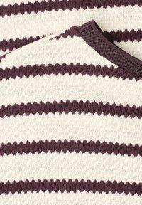 Nærbillede af en strikket sweater med vekslende horisontale creme og mørkelilla zigzagstriber og en ribbet mørkelilla halsudskæring.