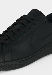 Μαύρο χαμηλό Nike sneaker με υφασμάτινη σόλα, ραμμένο λογότυπο Swoosh στο πλάι και μαύρα κορδόνια σε λευκό φόντο.