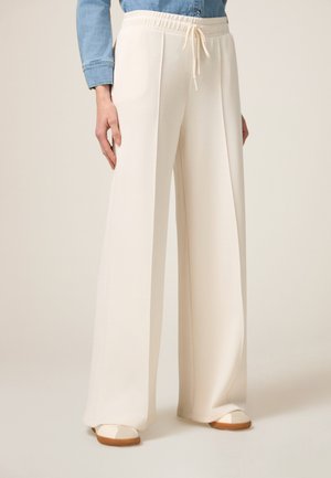 Pantaloni a gamba larga crema con una texture morbida, vita elastica e dettaglio con cordoncino, abbinati a scarpe basse chiare e una camicia di denim.
