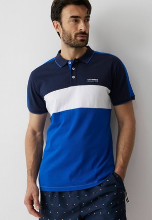 Mężczyzna w niebiesko-biało-granatowej koszulce polo z logo "Diverse Nautical Club", stoi z jedną ręką w kieszeni na jednolitym tle.
