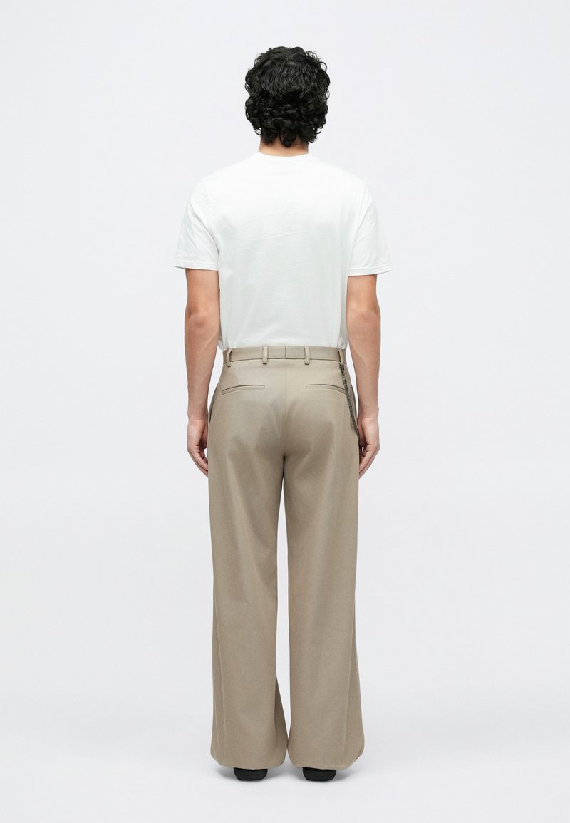 Man with curly black hair stands facing away, wearing a white t-shirt, beige wide-leg trousers with a belt, and black shoes on white background.