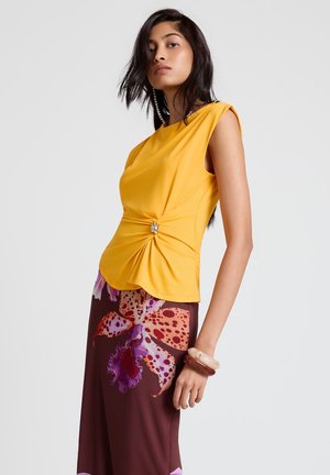 Femme portant un haut jaune sans manches avec un détail de boucle sur le côté et une jupe marron à grande impression florale rose et violette, debout de profil.