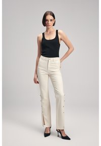Top senza maniche nero a costine con jeans bianchi a vita alta caratterizzati da fessure laterali e dettagli con bottoni. Il modello indossa tacchi a spillo neri.