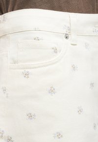 Κοντινό πλάνο λευκής φούστας από τζιν με μικρά μπλε και μπεζ λουλουδάτα σχέδια και μια μπροστινή τσέπη, φορεμένη με καφέ μπλούζα.