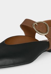 Chaussure plate noire pointue avec bride marron à la cheville et grande boucle ovale dorée sur fond blanc.