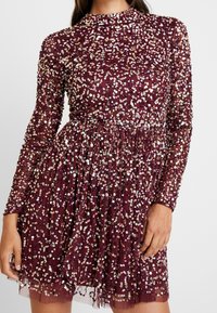 Kvinna i en långärmad, vinröd klänning täckt av iriserande paljetter med en figurnära midja och en rynkad kjol.