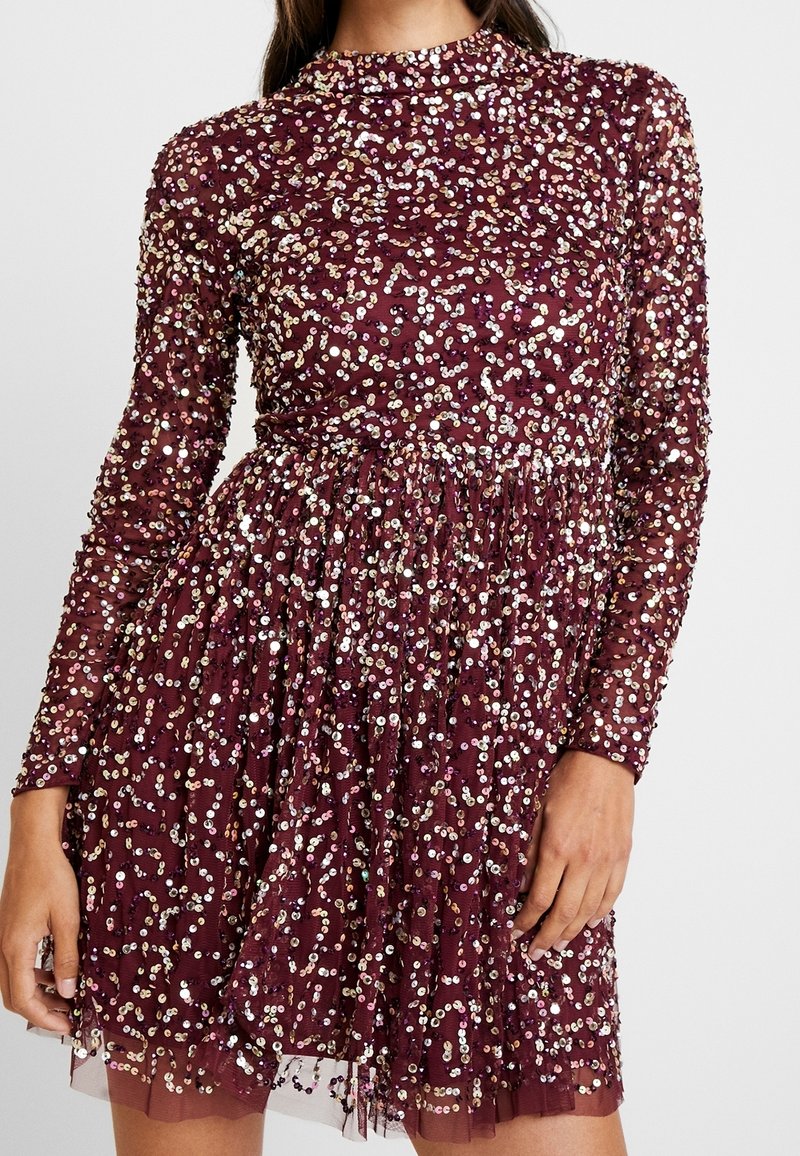 Kvinna i en långärmad, vinröd klänning täckt av iriserande paljetter med en figurnära midja och en rynkad kjol.