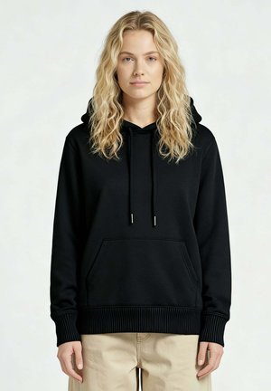 Donna bionda che indossa una felpa nera con cappuccio e tasca frontale e pantaloni beige, in piedi contro uno sfondo chiaro uniforme.