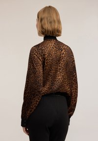 Bluzka w panterkę w brązowym i czarnym przezroczystym materiale. Posiada długie rękawy z mankietami na guziki oraz wysoki kołnierz, luźny krój.