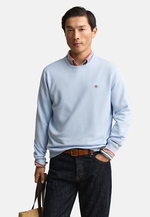 Man draagt een lichtblauwe trui over een gestreept overhemd, donkere spijkerbroek, bruine riem, houdt een beige draagtas vast op een witte achtergrond.