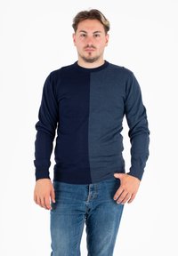 Mężczyzna z krótkimi włosami, ubrany w dwukolorowy granatowy sweter i niebieskie dżinsy, stoi na tle białym.