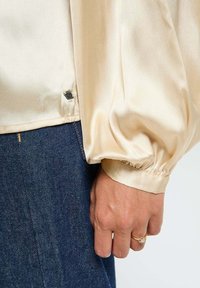 Blouse en satin crème à manches longues avec des poignets froncés, dotée d'un accent métallique à l'ourlet, associée à un jean en denim foncé.