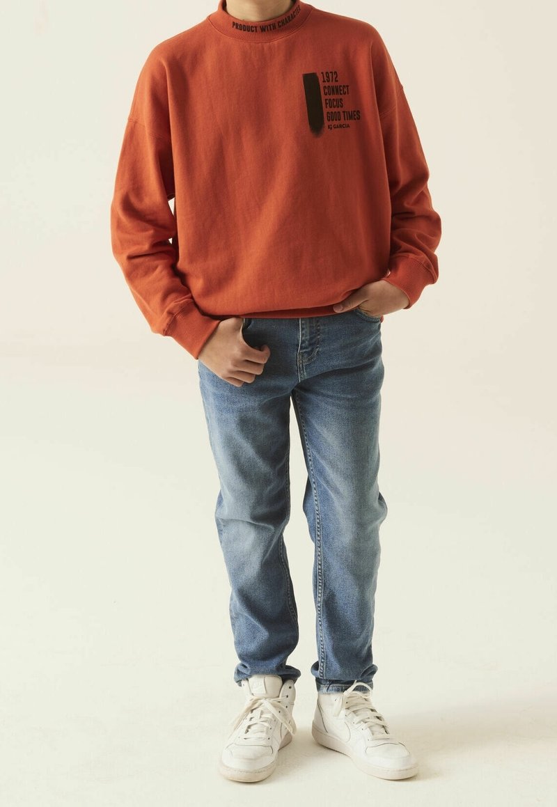 Sudadera naranja de cuello redondo con estampado negro, pantalones vaqueros de mezclilla azules y zapatillas blancas. Ajuste casual con detalles en los puños y el cuello de canalé.