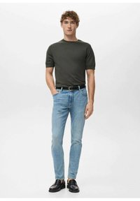 Tummanvihreä lyhythihainen t-paita yhdistettynä vaaleansinisiin kapealahkeisiin farkkuihin ja mustiin loafereihin, seisomassa neutraalilla taustalla.