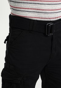 Černé cargo kalhoty s texturovanou tkaninou, vybavené širokým tkaným pásem, bočními kapsami a logem na levém stehně.