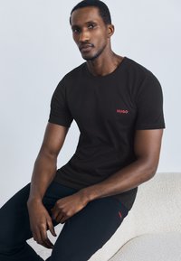 Černé tričko z bavlny s malým červeným logem "HUGO" na levé části hrudníku, doplněné texturovanými černými kalhotami. Model sedí na světlém povrchu.