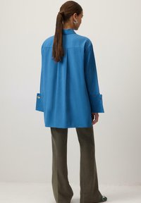 Sinine nööpidega särk tagumise voltiga, pikad varrukad mansettidega, kantud oliivsroheliste laia säärega pükstega. Sile kangas ja kohandatud disain.