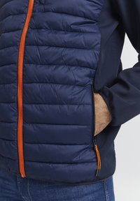Marinejakke med polstrede seksjoner, oransje glidelås, sidelomme og glatt stoff på ermene. Strukturelt quiltet design gir dimensjon.