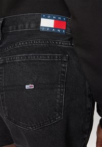 Svarta denimjeans med bakficka som har en liten Tommy Hilfiger-flagga och en större logotypapplikation nära midjan på svarta kläder.
