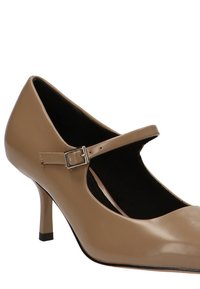 Zapato beige de charol tipo Mary Jane con tacón bajo, correa en el tobillo y forro interior negro. Presenta una punta redondeada y una silueta elegante.