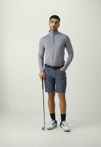 Vyras stovi su golfu, vilkintis pilką ilgomis rankovėmis užtraukiamą marškinėlį ir tamsiai mėlynus šortus su pakartotiniais "Calvin Klein" užrašais.