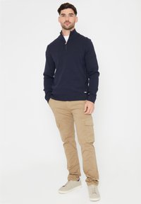 Marineblauwe zip-nektrui van een zacht materiaal, gecombineerd met beige cargobroeken met zijzakken en een taps toelopende pasvorm. Lichte sneakers.