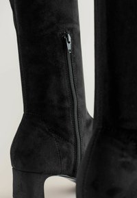 Bottes noires en daim montantes avec fermeture éclair sur le côté et talon haut effilé. Texture lisse avec des détails de couture minimaux.