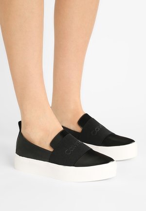 Fødder iført sorte slip-on sneakers med tykke hvide såler og brede sorte elastikbånd ovenpå med subtil "Calvin Klein" branding.