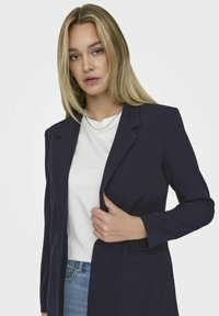 Jūras zilas piegriezuma žakete ar izgrieztu apkakli un vienu pogu, ar šaurām garām piedurknēm un gludu tekstūru.