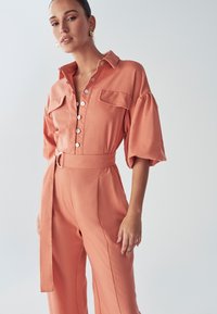 Mujer vestida con un mono coral de botones, con cinturón ancho y mangas abullonadas, de pie contra un fondo claro y liso.