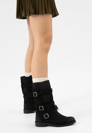 Benen met witte sokken en zwarte laarzen tot halverwege de kuit, elk met drie zilveren gespen, gecombineerd met een bruine geplooide rok.