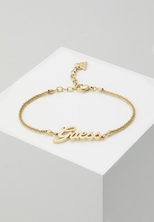 Bracelet chaîne en or avec inscription « Guess » au centre et fermoir ajustable, présenté sur une surface blanche inclinée.