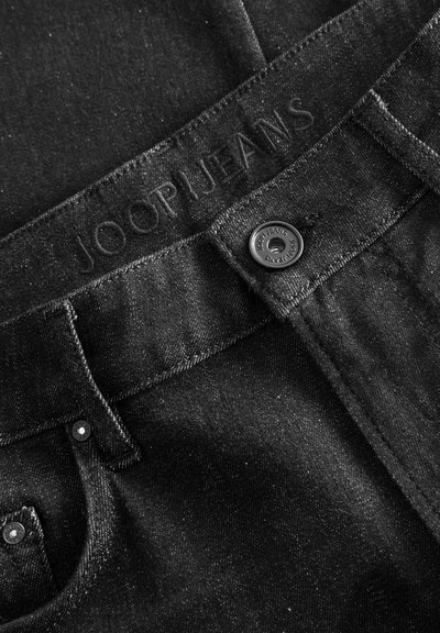 Jean en denim noir avec une finition texturée, logo embossé à la taille et fermeture à bouton en métal avec logo.