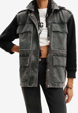 Persona con una chaqueta vaquera negra y gris con varios bolsillos con solapa sobre una camiseta corta blanca con gráfico y pantalones negros.