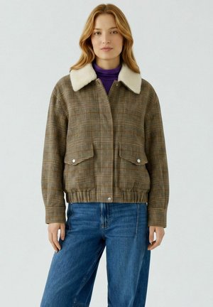 Femme portant une veste marron à carreaux avec un col en fourrure blanche par-dessus un haut violet et un jean bleu, debout devant un fond clair uni.
