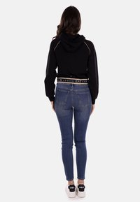 Felpa nera corta con cappuccio, presenta una banda con il logo in vita. Indossata con jeans skinny blu e sneakers bianche con dettagli neri.