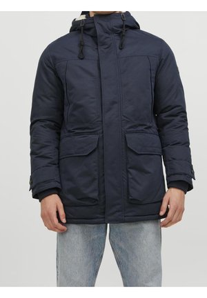 Mann trägt marineblauen Kapuzen-Wintermantel mit vorderen Klappentaschen und hellblauen Jeans vor einfachem Hintergrund.