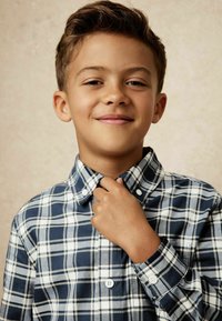 Camisa de cuadros azul y blanca, con cuello, de diseño ajustado y mangas largas, mostrada en un niño que está ajustándose el cuello.