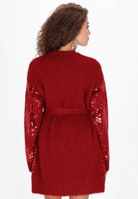 Czerwony dzianinowy kardigan z długimi rękawami, zdobiony cekinami, z paskiem w talii i teksturowanym wykończeniem. Zaprojektowany w luźnym fasonie.