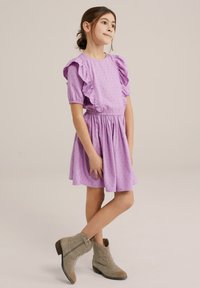 Ljubicasta pamučna haljina s napučenim rukavima i detaljima od ruffles-a, sužena u struku, uparena sa bež čizmama do članka. Glatka tekstura s suptilnim uzorcima.