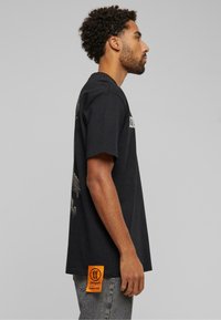 Sort bomulds T-shirt med grafik på ryggen og en orange label på siden. Korte ærmer og afslappet pasform.