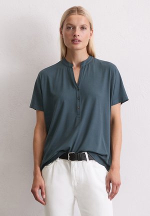 Blouse à manches courtes teal avec un col montant, patte de boutonnage à six boutons et coupe décontractée, associée à un pantalon blanc et une ceinture noire.
