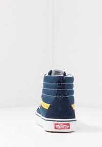 Vans vysoké tenisky v námořnické modré semišové kůži se žlutým bočním pruhem, bílou gumovou podrážkou a výrazným červeným logem na patě. Zpevněné švy.