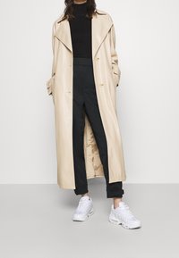 Béžový dlouhý trenčkot s širokým límcem a zlatými knoflíky, nošený přes černý top a černé kalhoty. Bílé tenisky dokončují outfit.