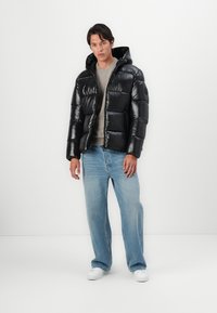 Black puffer jacket with shiny texture, hood, and quilted sections, worn over a grey sweater and light blue jeans. White sneakers are visible.