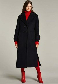 Cappotto lungo in lana nera con chiusura doppiopetto, ampi revers e tasche laterali. Abbinato a un dolcevita rosso a costine e collant rossi.